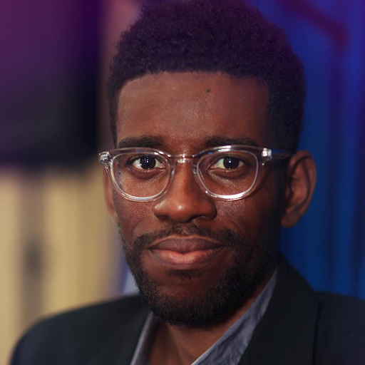 A man with short dark hair and clear-framed glasses, wearing a dark blazer over a grey shirt against a blue background.