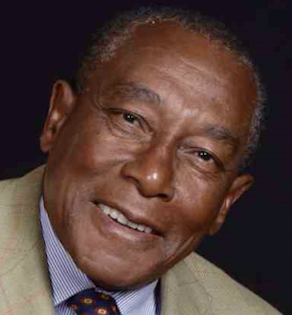 A headshot of a person smiling, wearing a light tan blazer, a striped shirt, and a patterned tie against a black background.