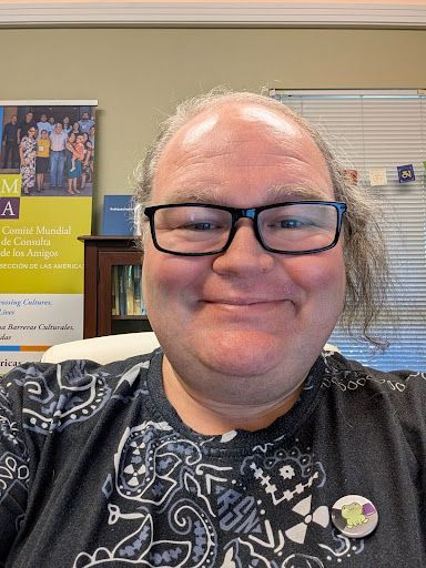 A person with long hair and glasses smiles in an office setting with a poster in the background.