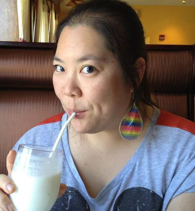 A person with a rainbow teardrop earring sips a white drink through a straw in a booth setting.