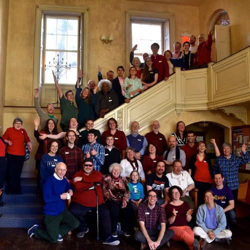 A large group poses on and around a grand staircase inside a bright, historic building with tall windows.