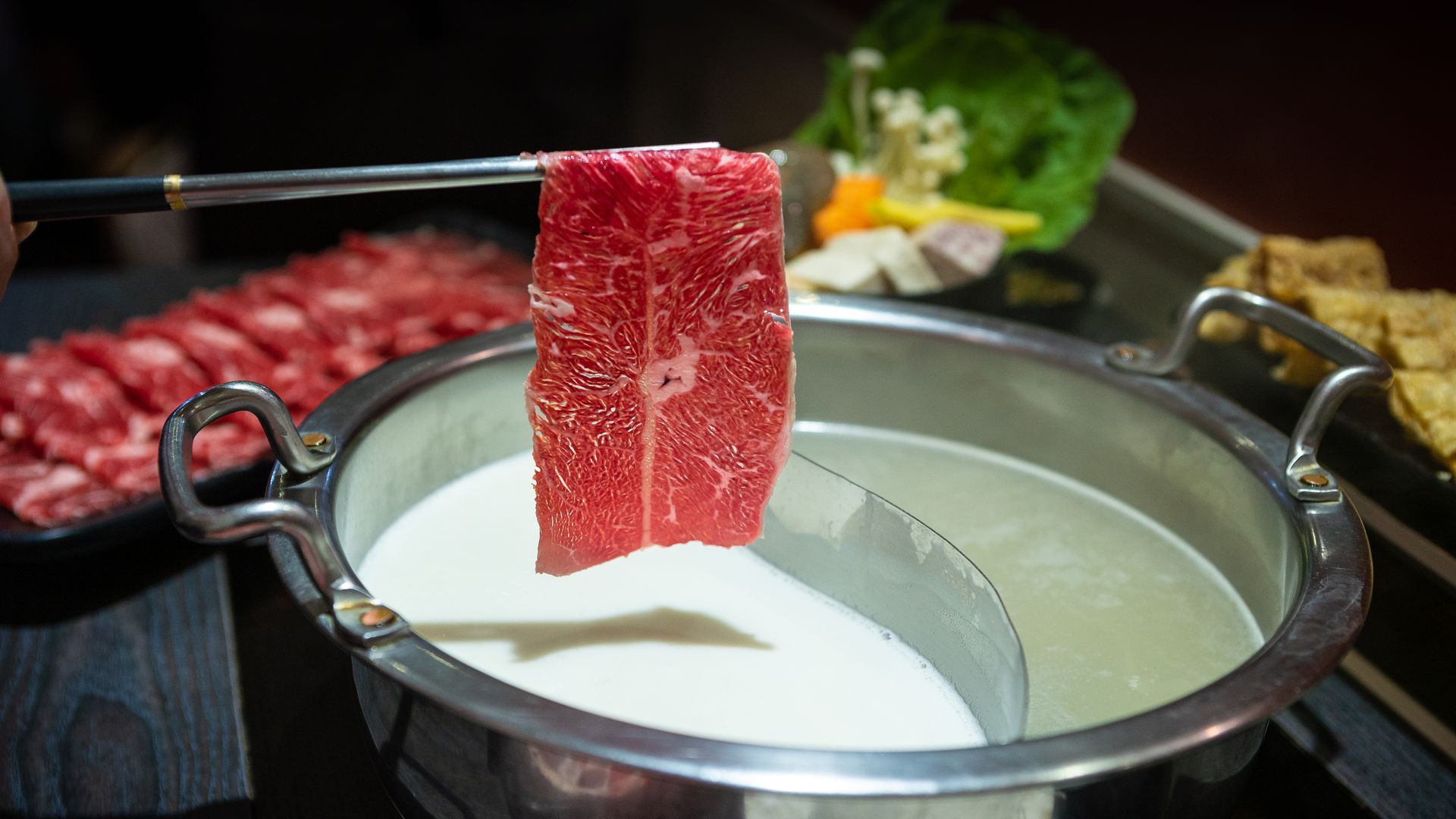 JPOT Japanese hot pot metal chopsticks holding raw sliced meat over hot pot broth, preparing to cook