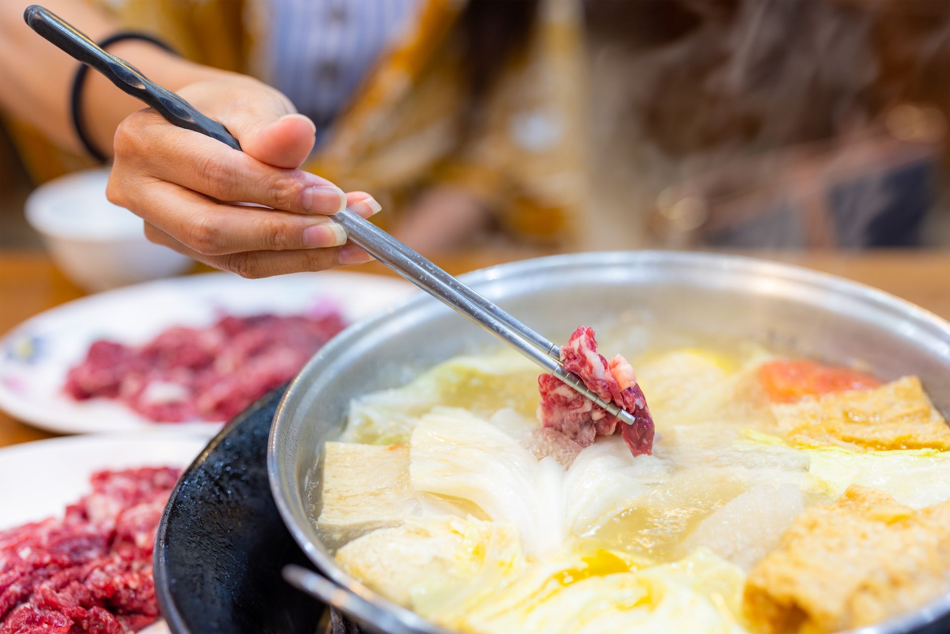 JPOT Japanese hot pot person dipping raw meat into hot pot broth to cook with raw meat nearby and veggies and tofu in broth