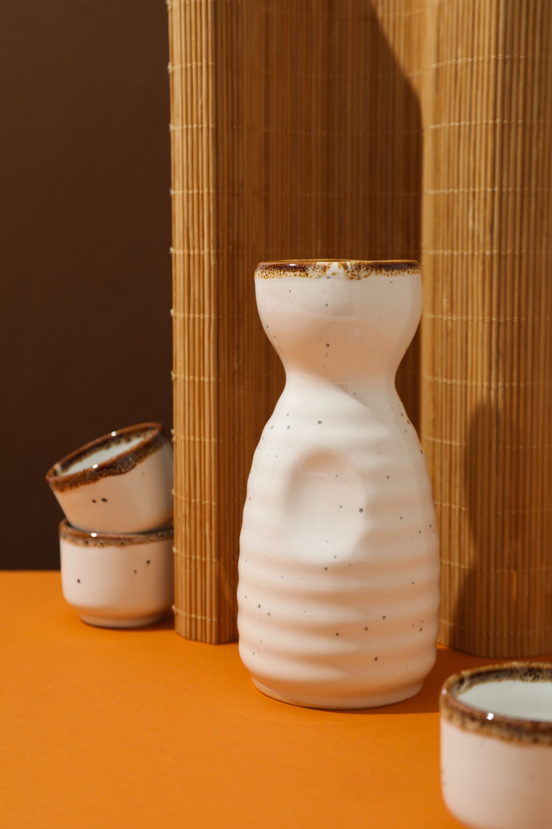 JPOT Sake white and brown rimmed ceramic pitcher with two matching ceramic cups in the background with bamboo mat decor