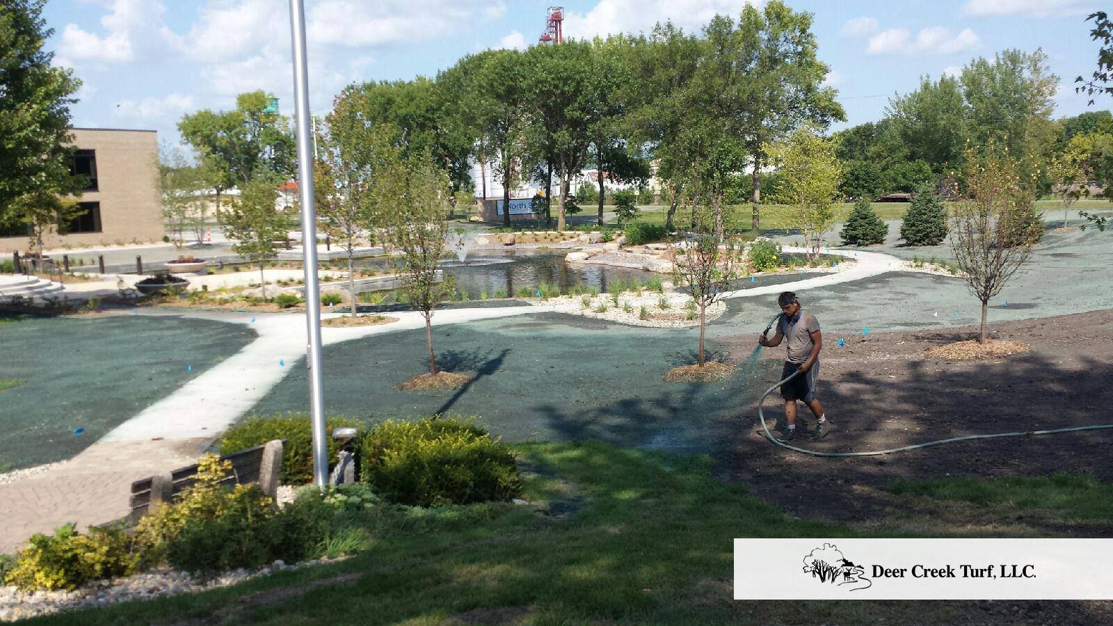 A landscape worker sprays hydromulch over a newly seeded area near a park pond on a sunny day.