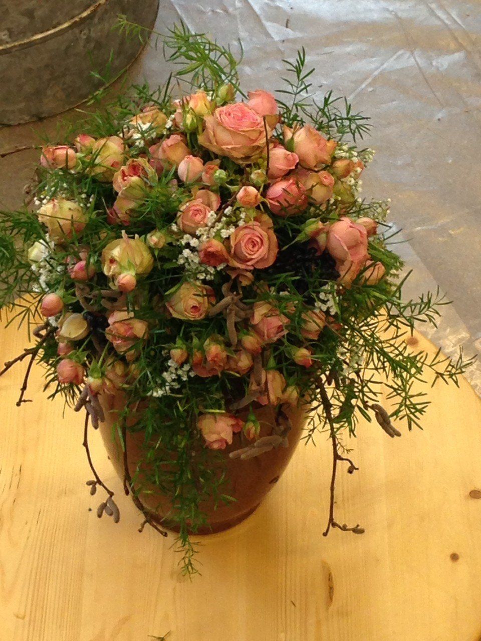 Auf einem Holztisch steht eine Vase mit rosa Rosen und Grünpflanzen.