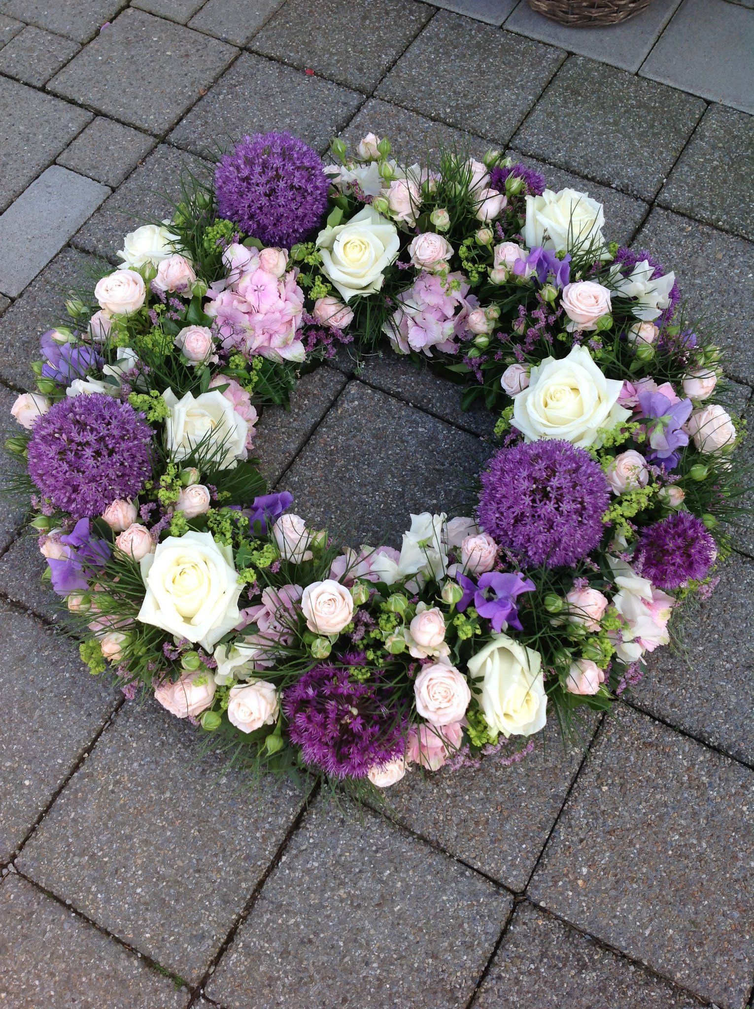 Auf einem gepflasterten Gehweg liegt ein Kranz aus violetten und weißen Blumen.