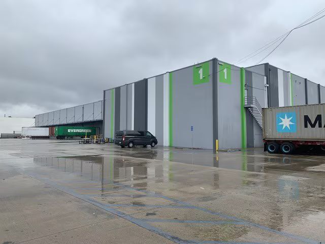 Warehouse exterior with loading docks, semi-trucks, and a black van on a wet, overcast day.