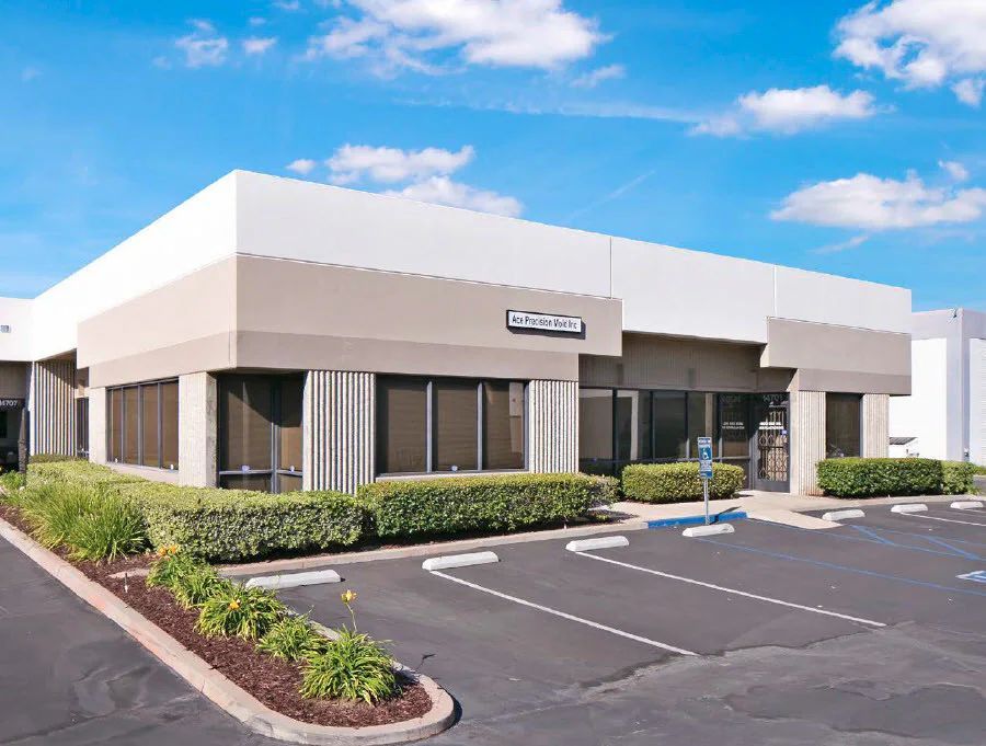 Commercial building with tan and white facade, windows, and parking spaces. Blue sky and greenery.