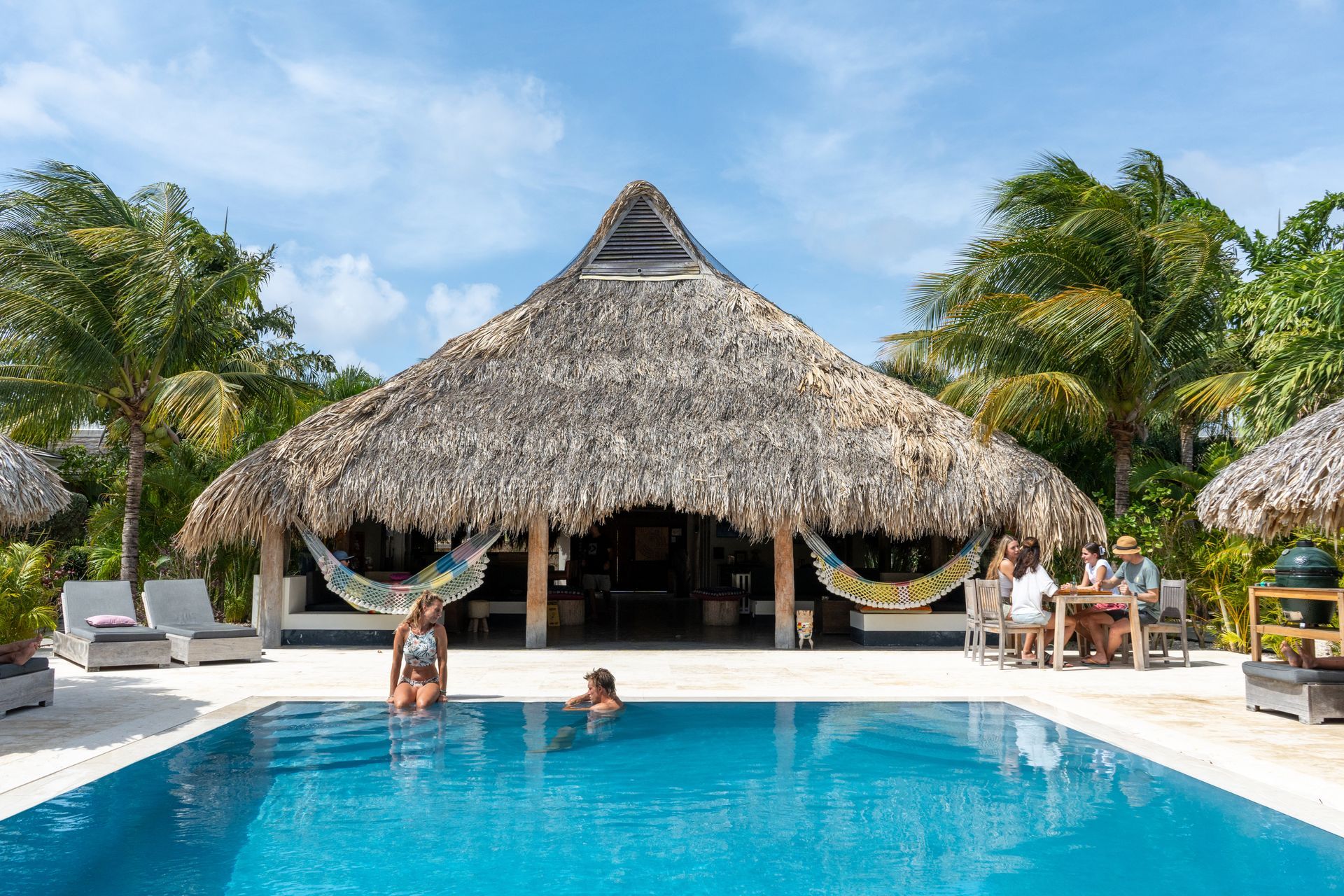 A man and a woman are swimming in a pool in front of a thatched hut.