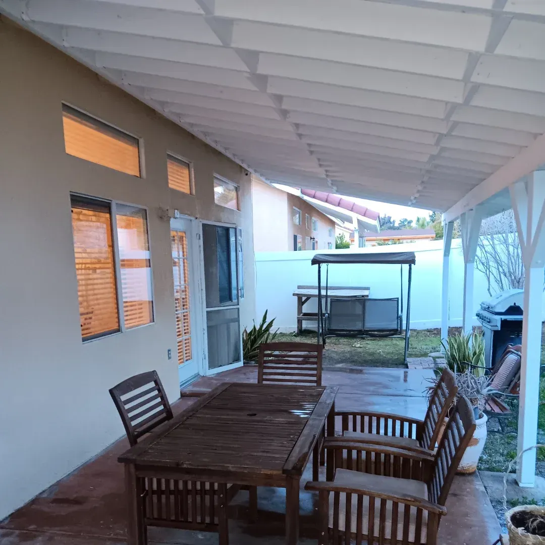 Covered patio with wooden table and chairs beside a house, with a small pool or hot tub in the background