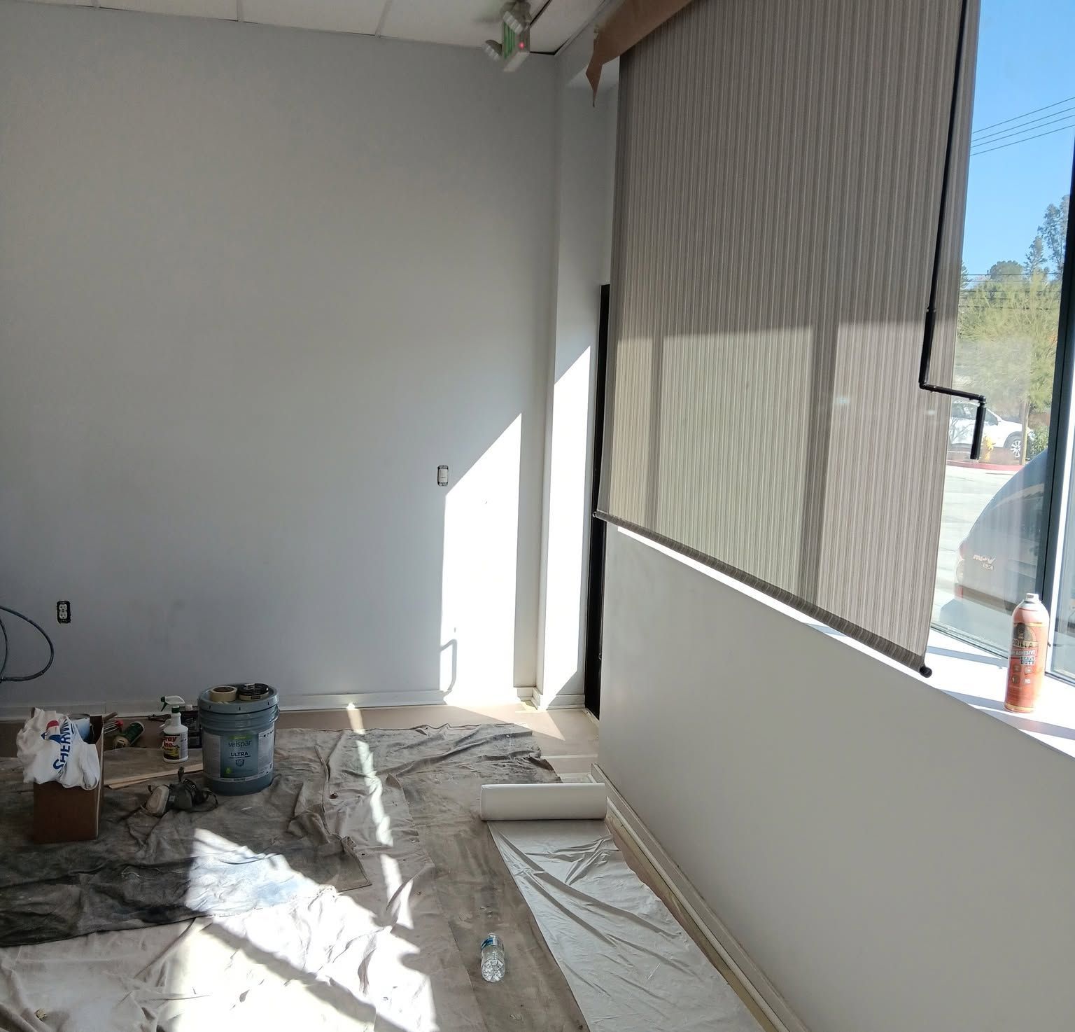 Sunlit room under renovation with white walls, covered floor, paint buckets, and a large window blind.