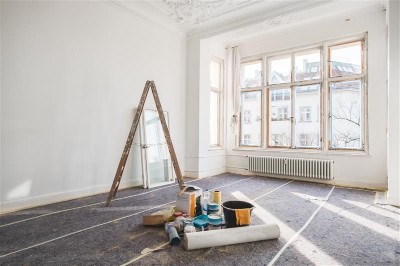 Bright empty room with a ladder, paint supplies, and tools on a gray floor near a large window.