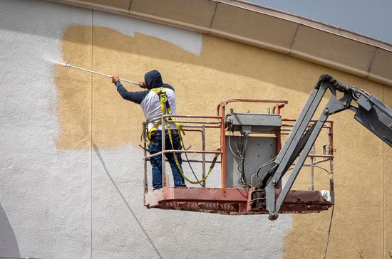 Worker on a lift spraying tan paint onto a building wall.