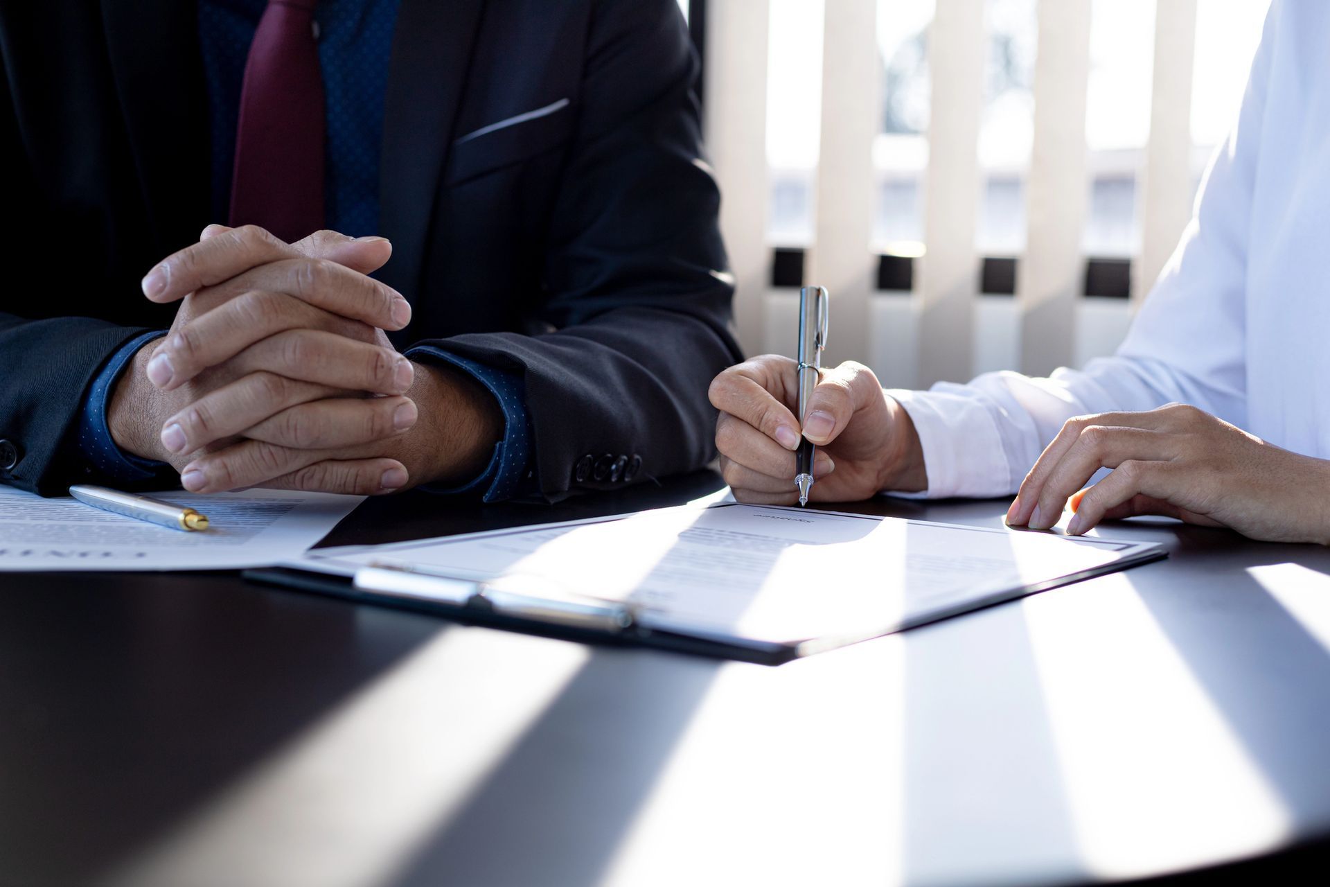 Due persone siedono a un tavolo e stanno esaminando dei documenti, una con le mani giunte e l'altra intenta a scrivere con una penna.