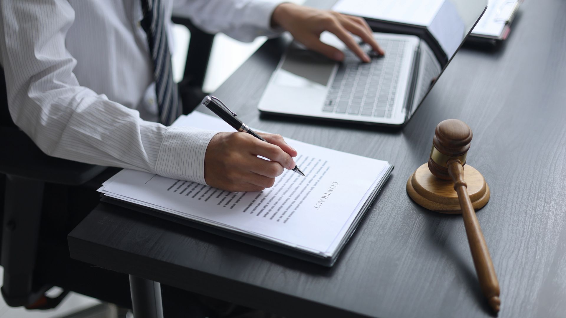 Una persona in abiti professionali firma un documento a una scrivania, con un computer portatile e un martelletto di legno nelle vicinanze.