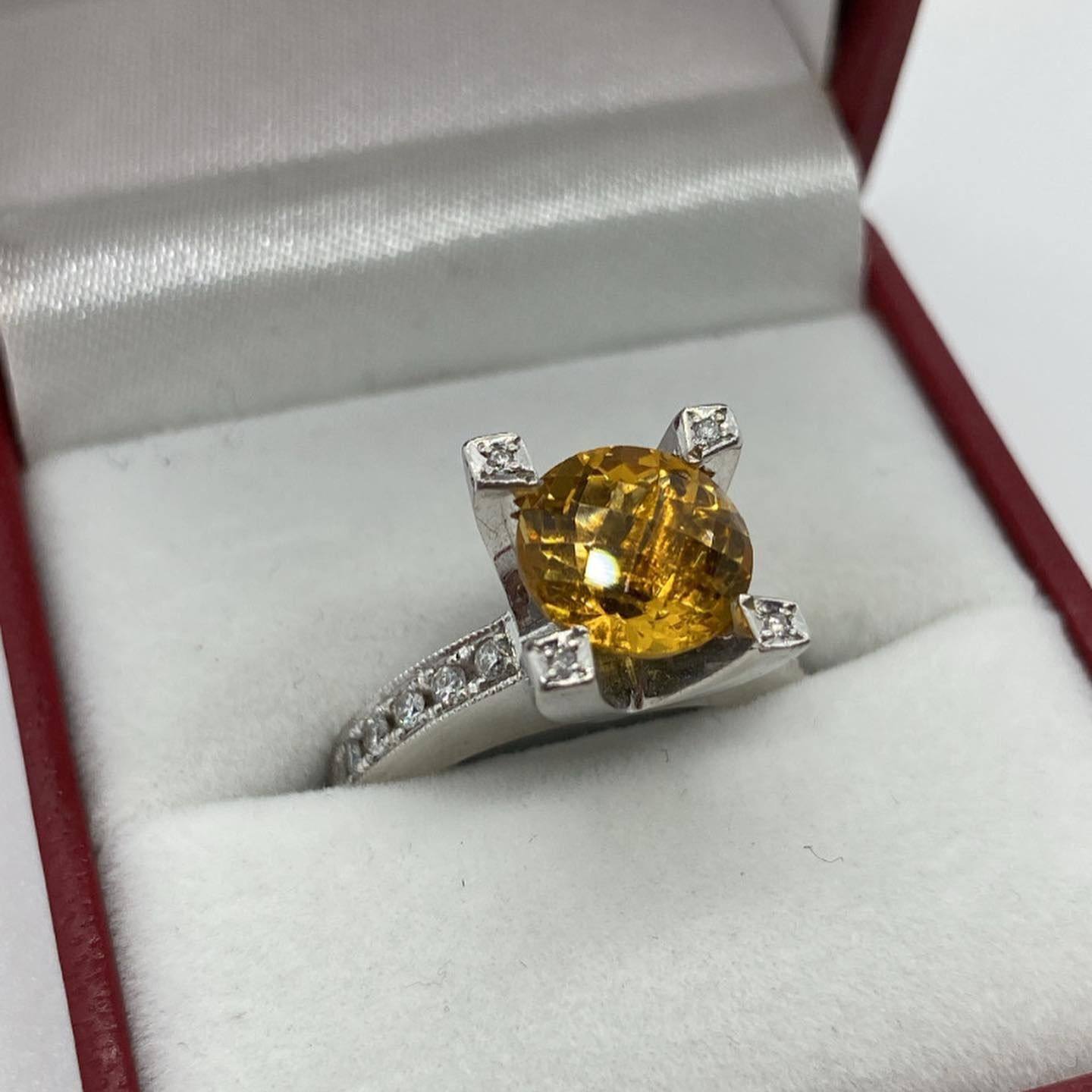 A silver ring with a round, yellow gemstone and small diamonds on the band, displayed in an open red jewelry box.