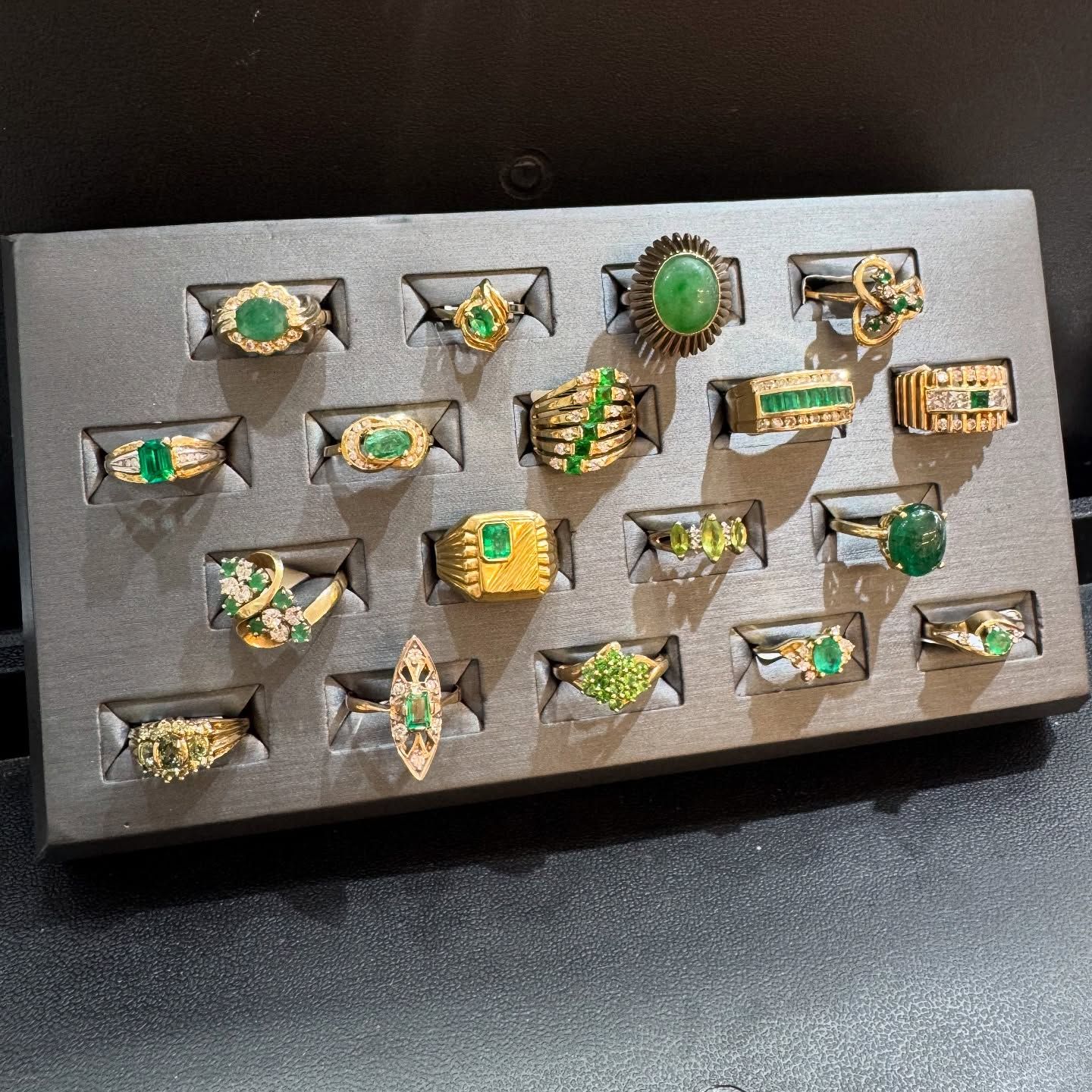 A display tray holding 16 gold rings featuring various green gemstones set in a gray foam organizer.