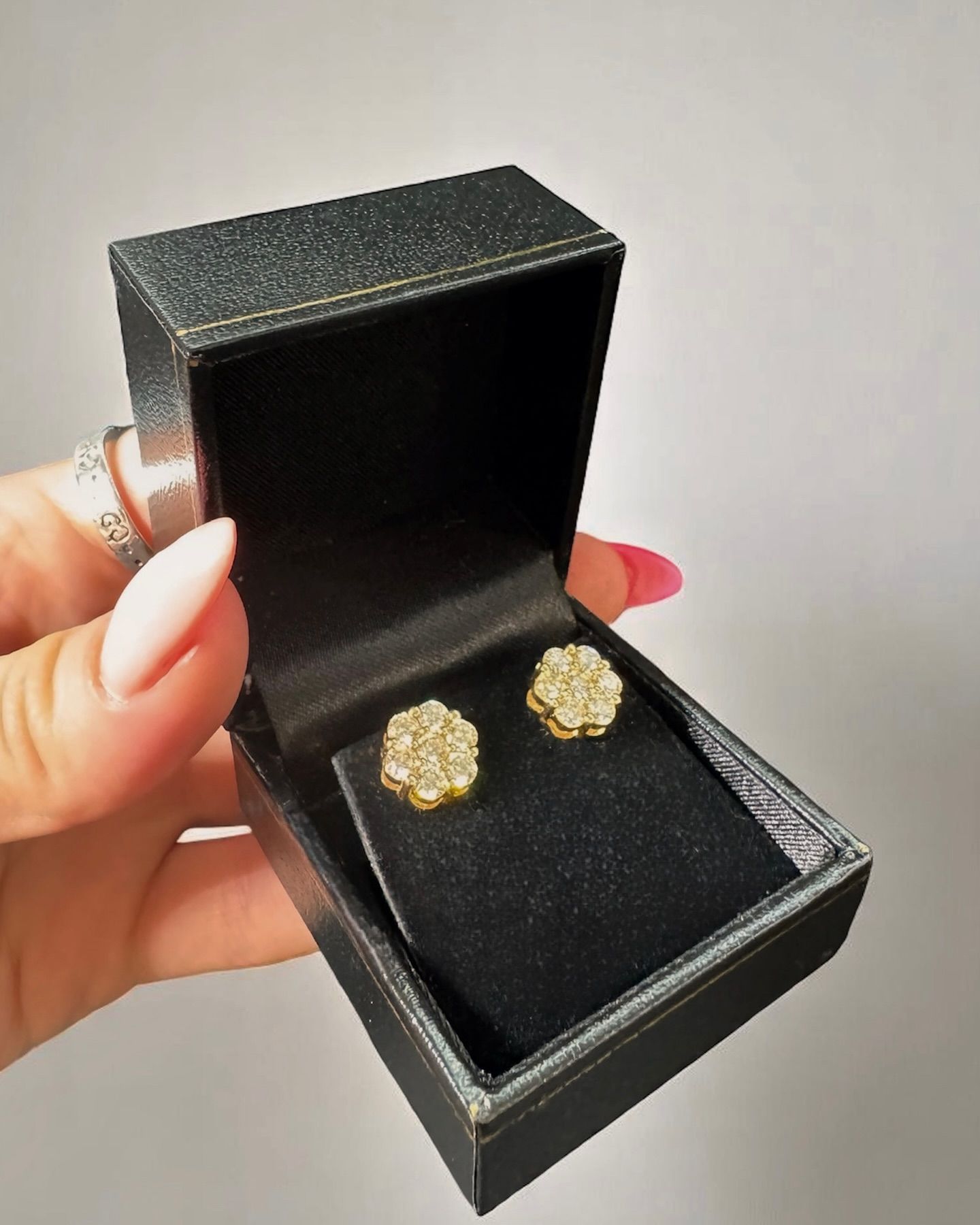 A hand holds an open black jewelry box containing a pair of gold floral-shaped stud earrings with embedded stones.