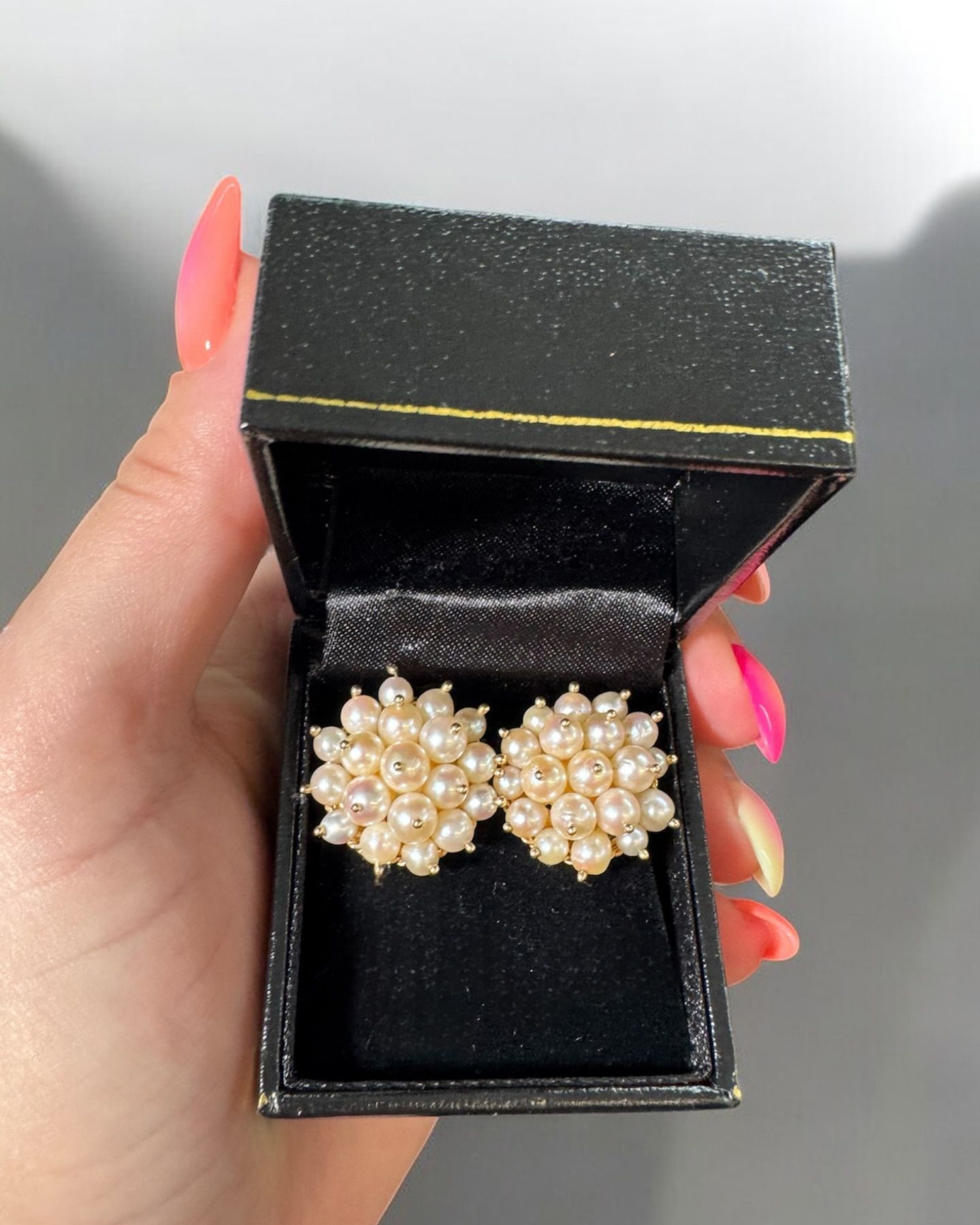 A hand holds an open black jewelry box containing a pair of vintage-style floral cluster pearl earrings.