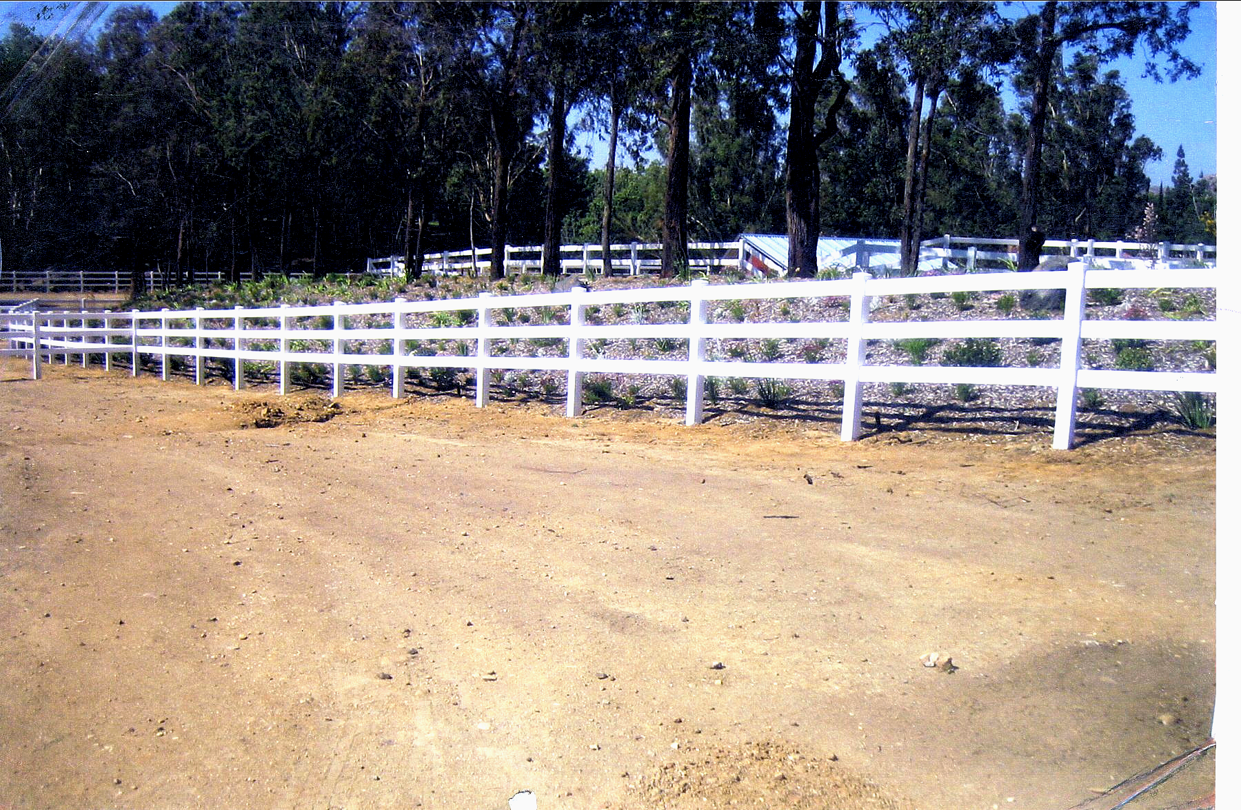 A white fence surrounds a dirt field with trees in the background