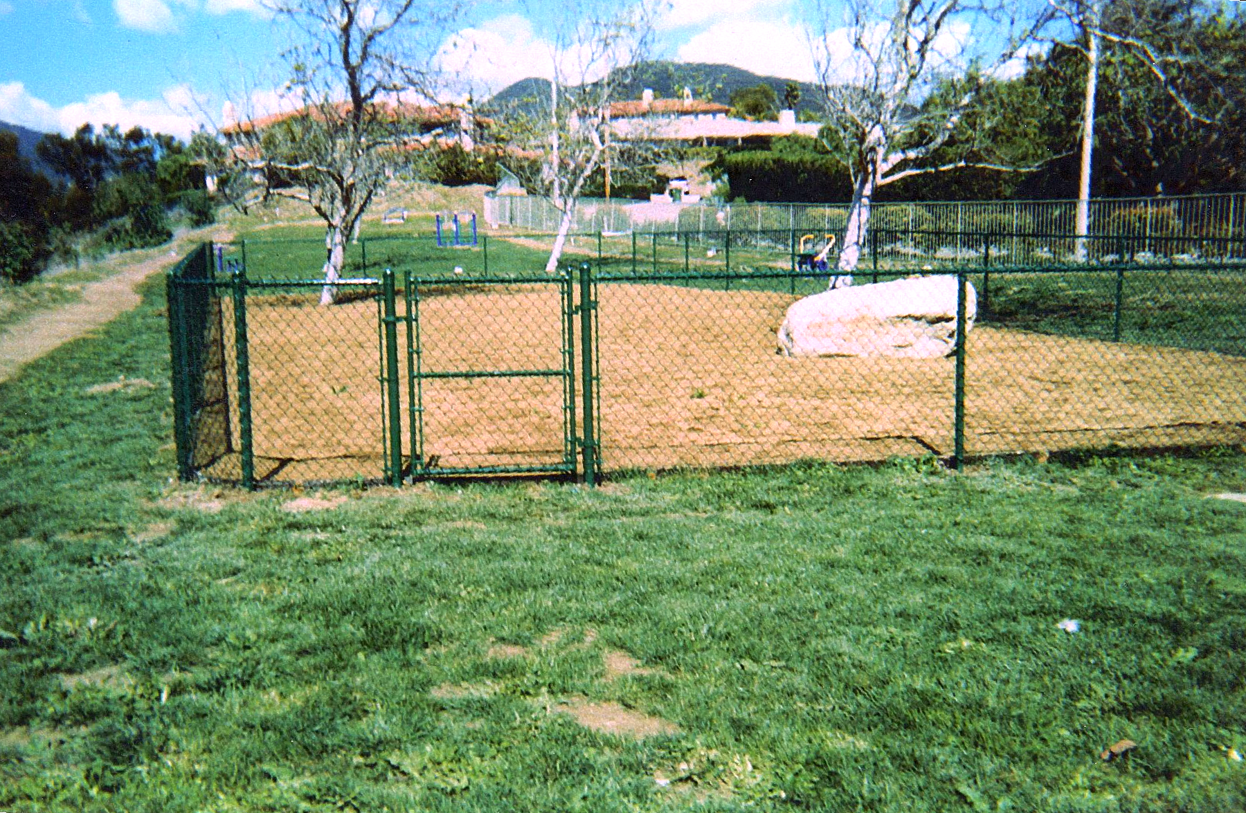 A chain link fence surrounds a dirt field in a park.