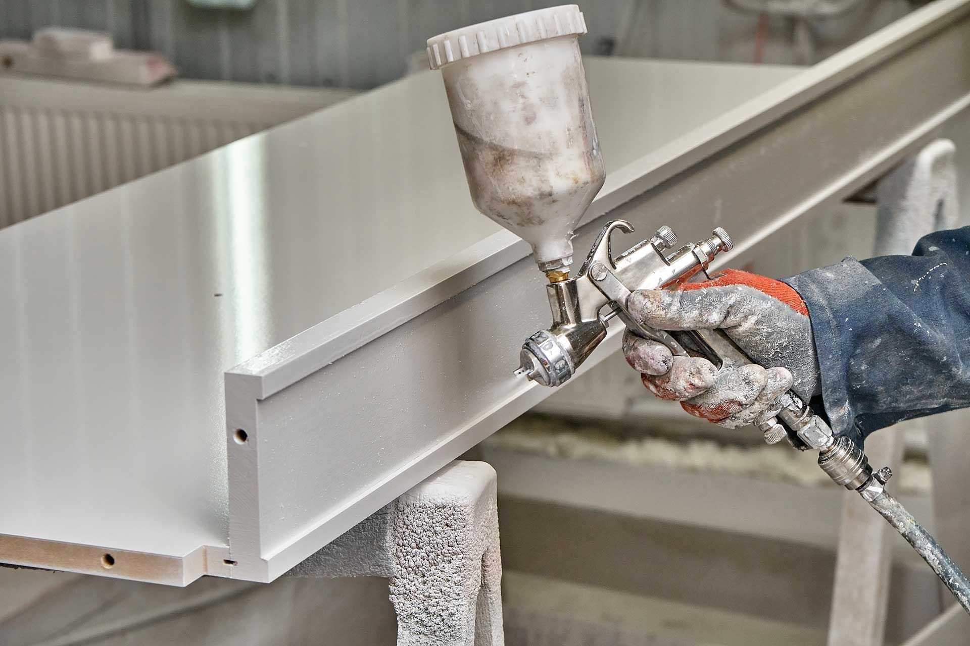 Person sprays white paint on a wooden shelf with a paint sprayer in a workshop.