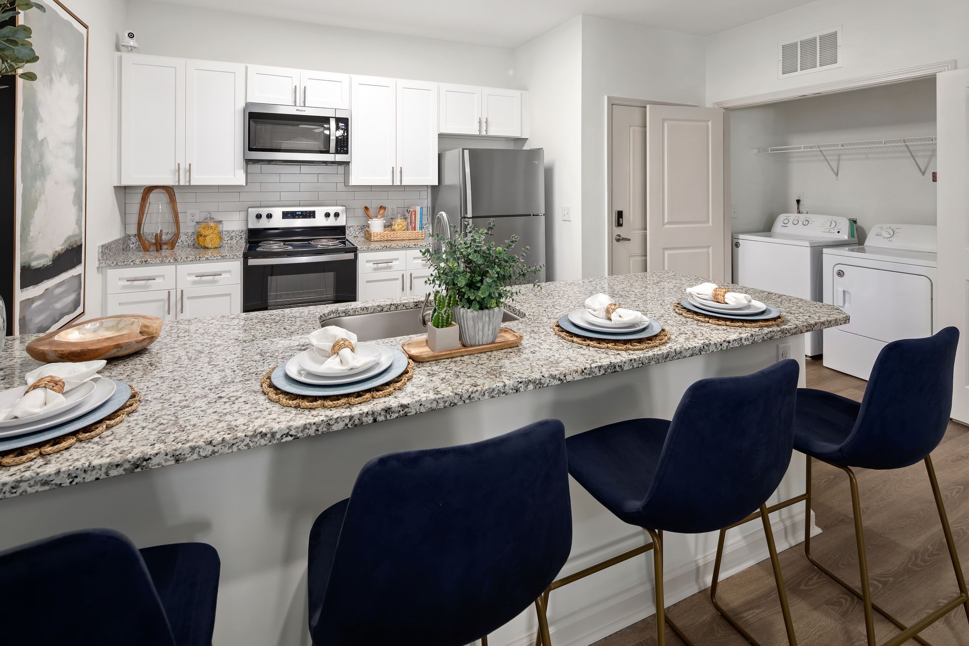 Modern kitchen with granite island, navy bar stools, and white cabinetry. Laundry in background.