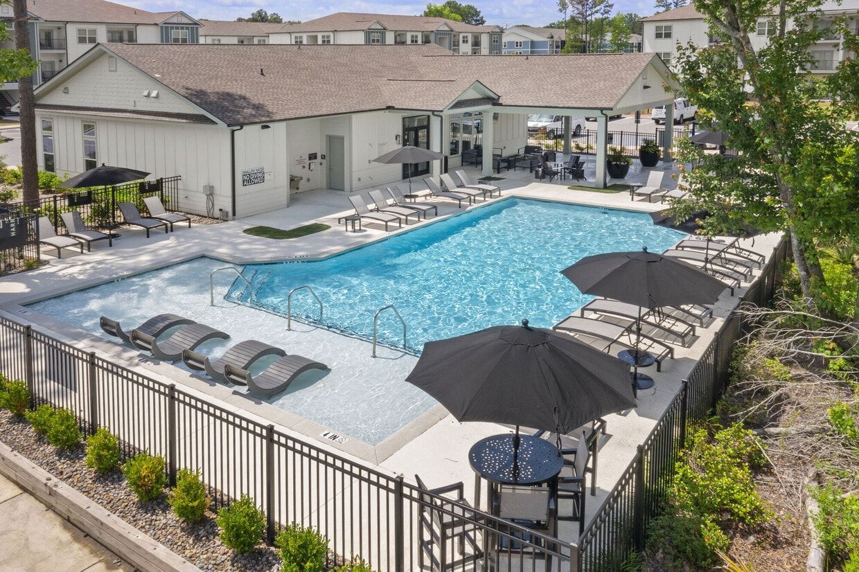 Pool area with lounge chairs, tables with umbrellas, and a white building in the background.
