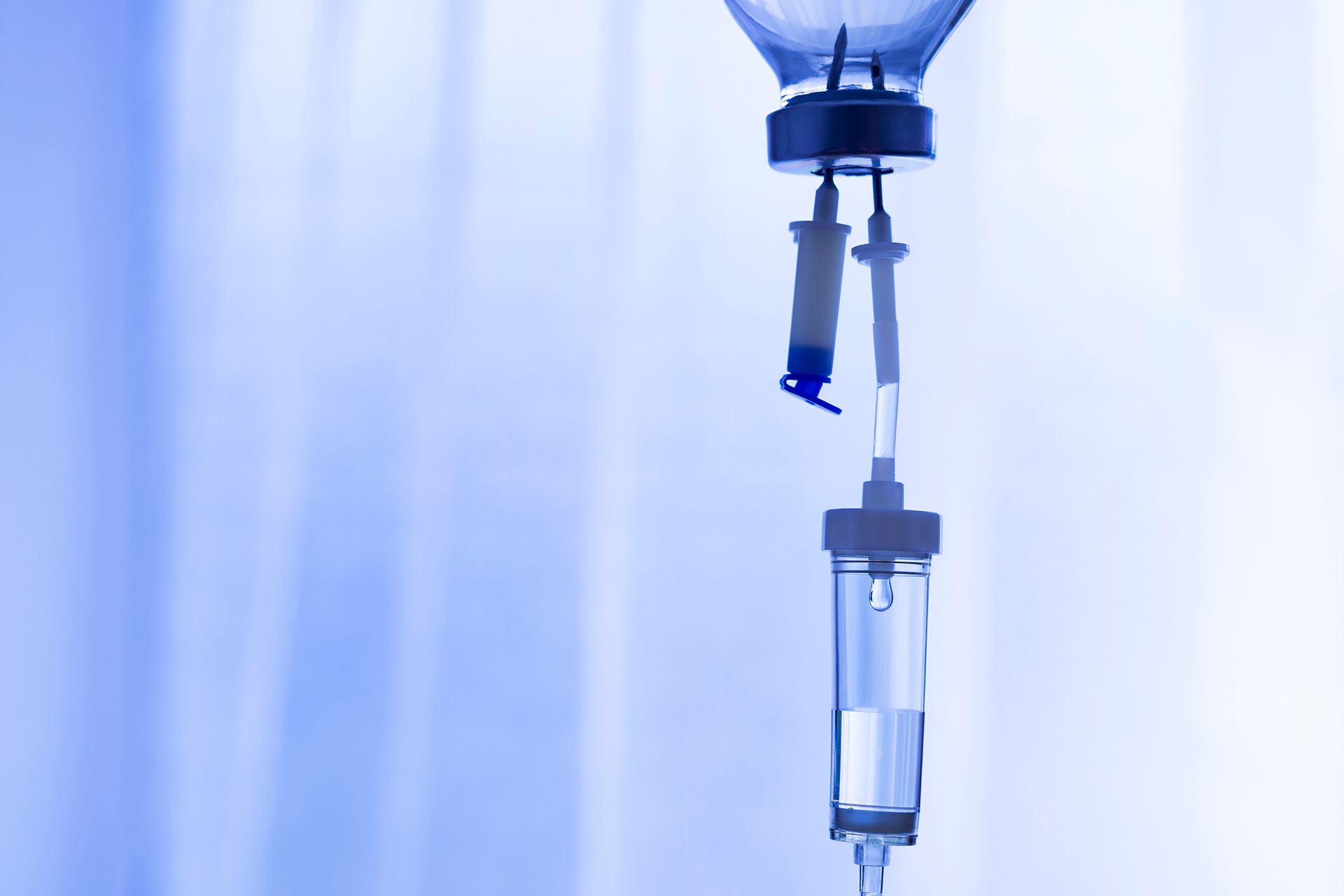 A blue-toned close-up of an IV drip bag and fluid chamber hanging against a blurred background.