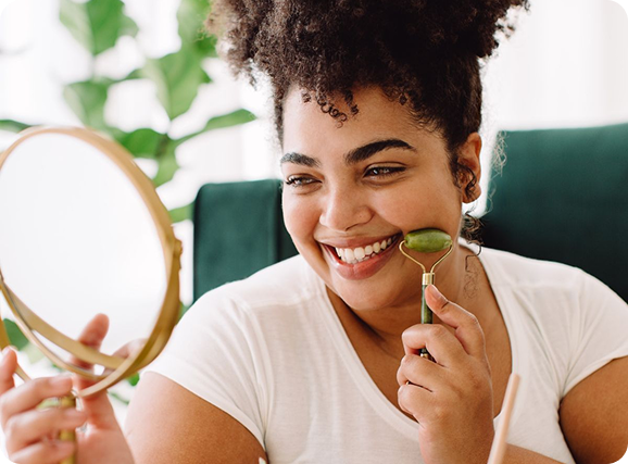 A woman is smiling while using a jade roller on her face.