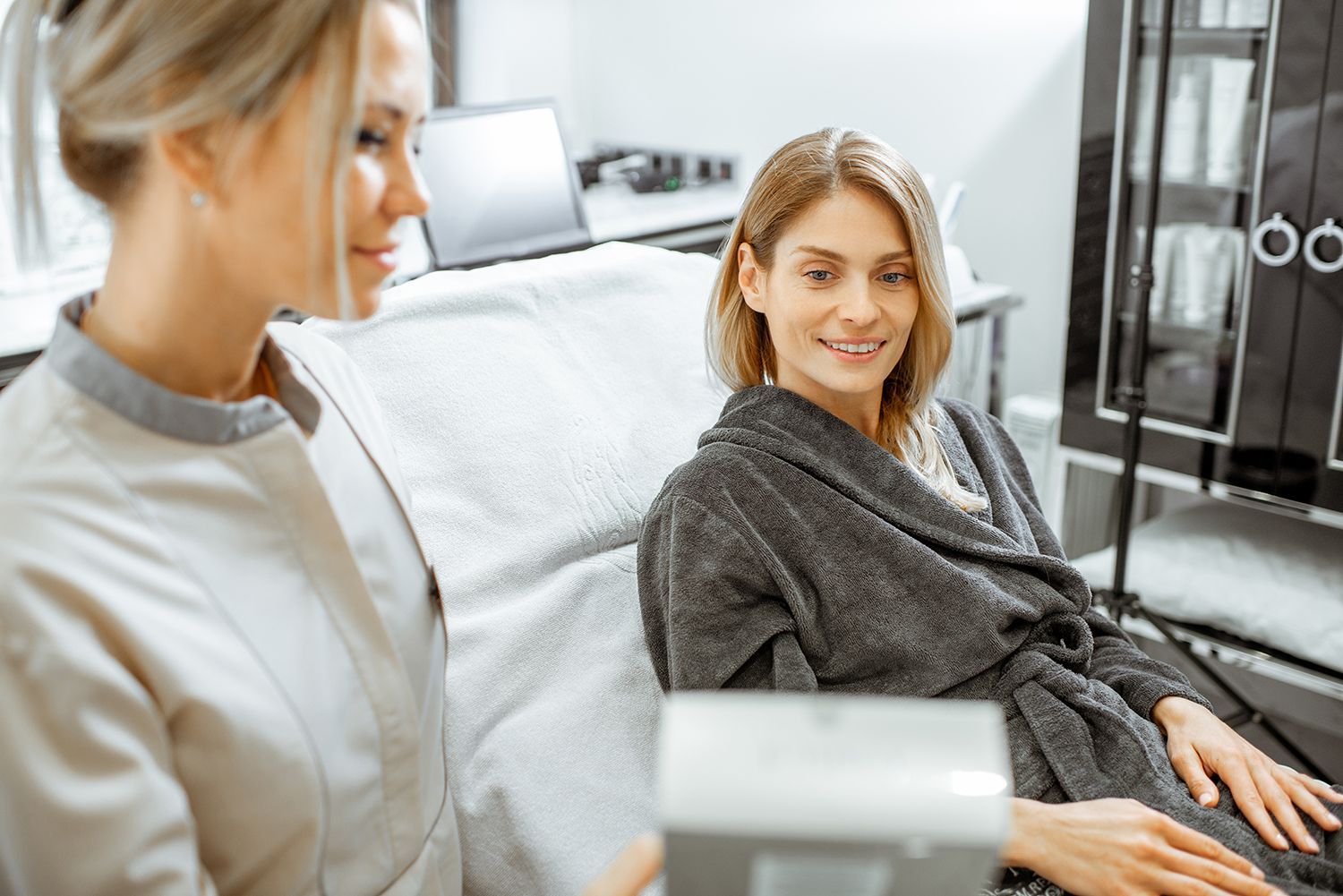 A beauty specialist in a uniform explains a product to a client wearing a dark robe in a bright, modern spa treatment room.