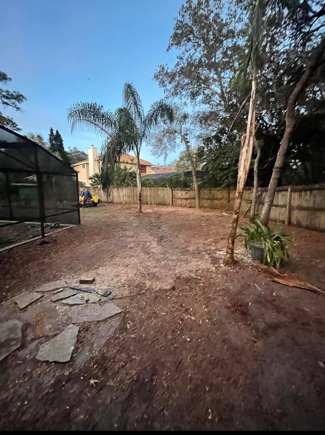 Backyard with dirt ground, wooden fence, screen enclosure, trees, and a house in the distance.