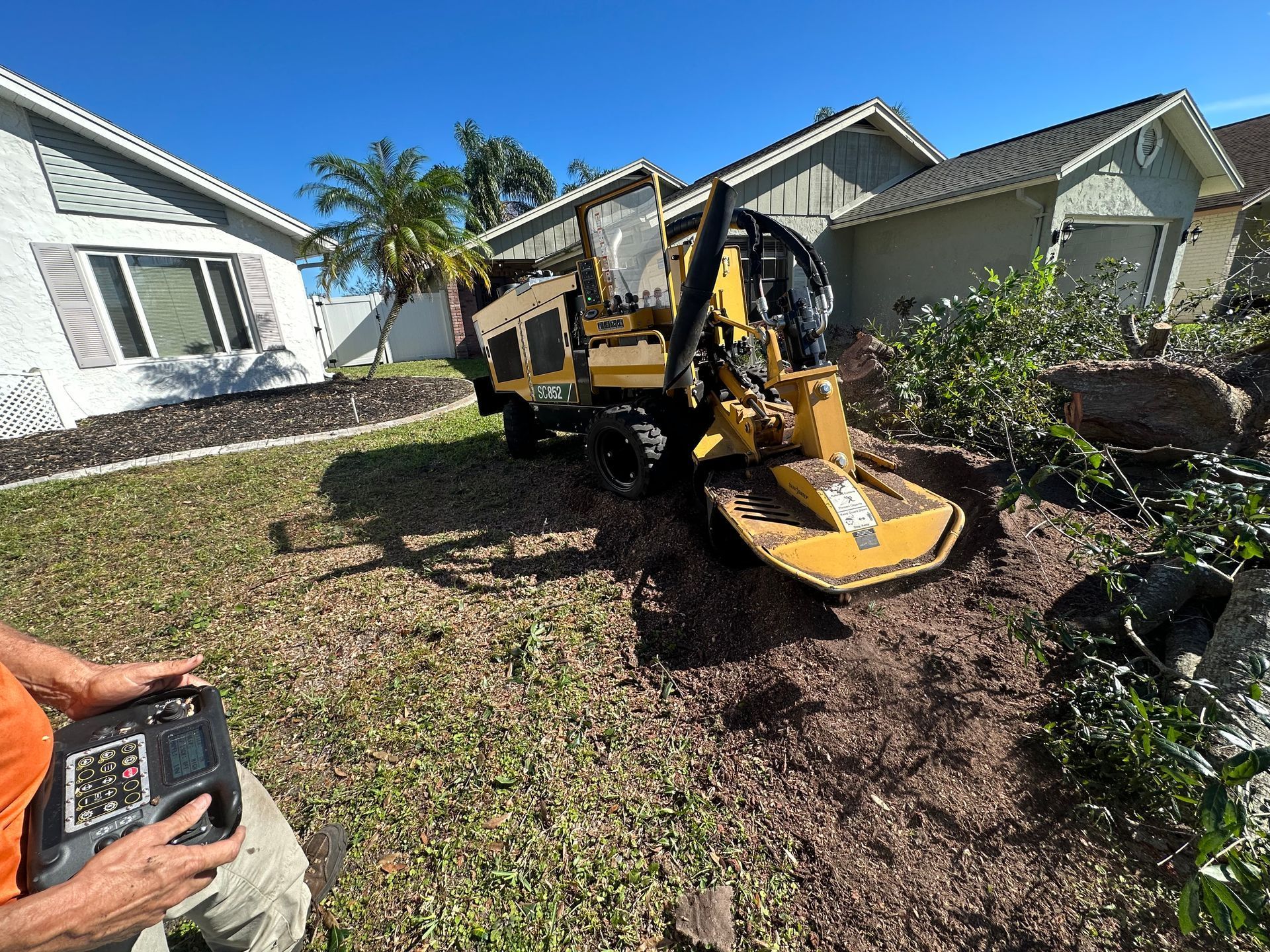 A yellow stump grinder on a lawn, controlled by a person. Houses in the background.