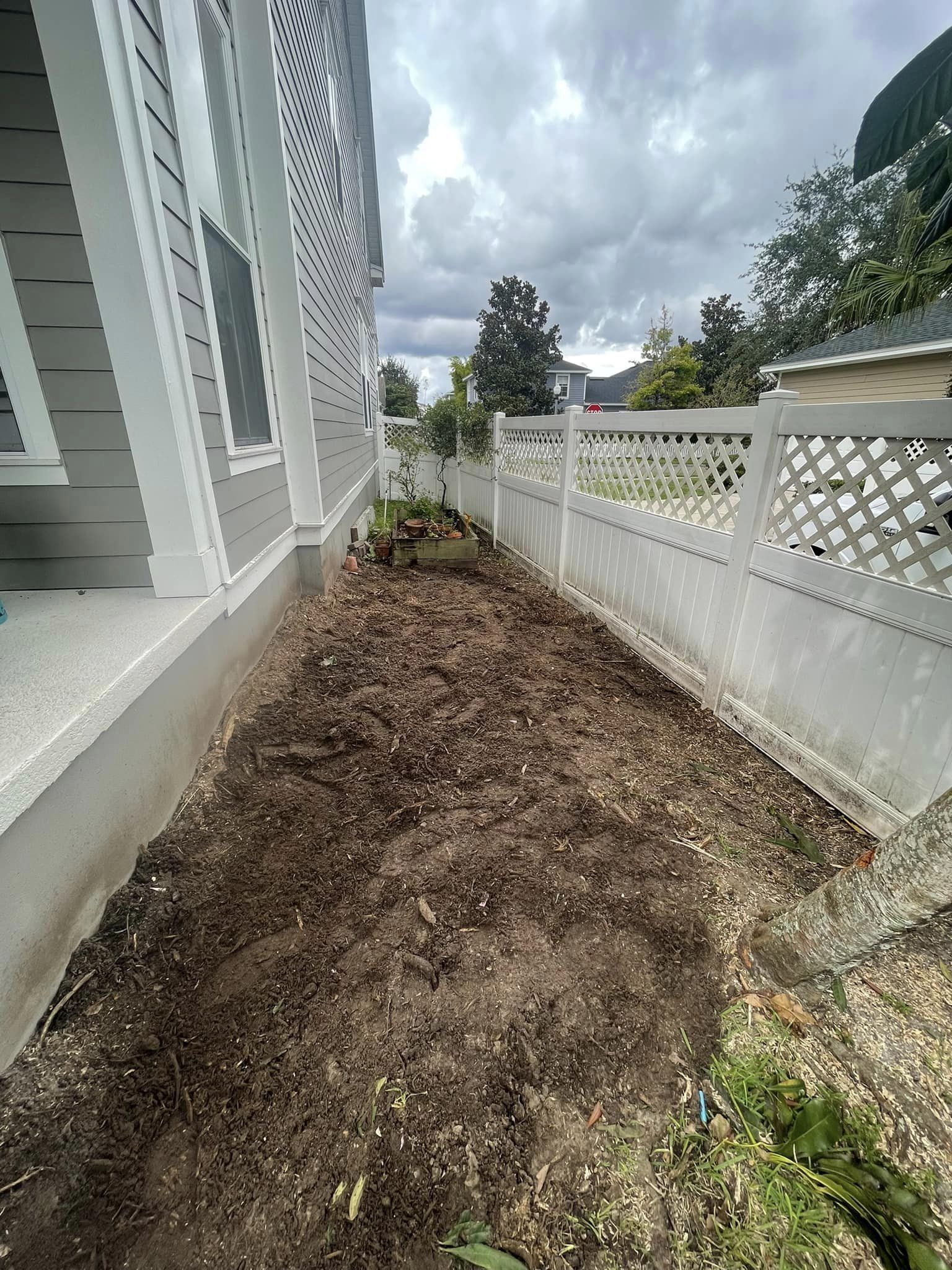 A long, narrow garden bed next to a house and fence, filled with dark soil under a cloudy sky.