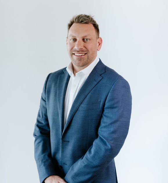 Man in blue blazer and white shirt smiles against a white backdrop.
