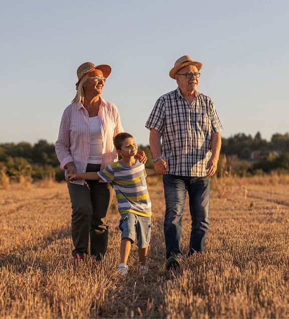 Grandparents and child walking in a field, holding hands, wearing hats. Golden sunlight.