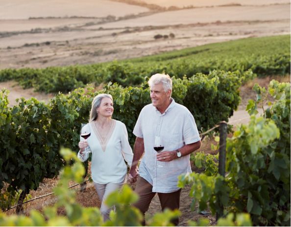 Couple holding hands, smiling, walking through a vineyard while enjoying wine.