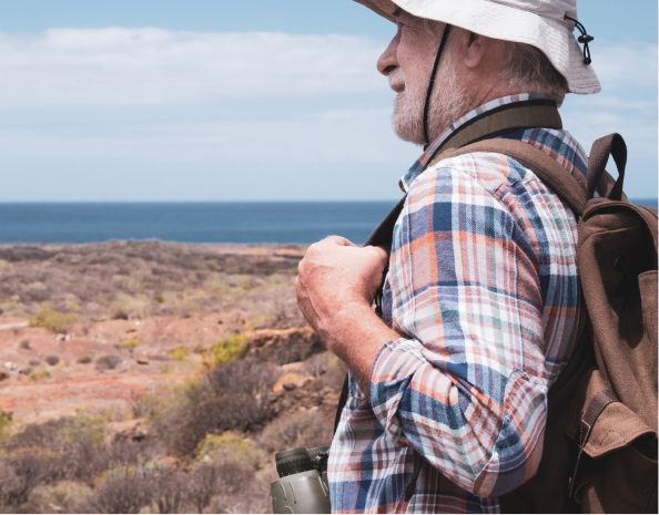 Man in plaid shirt and hat with backpack, looking at a coastal landscape with binoculars.