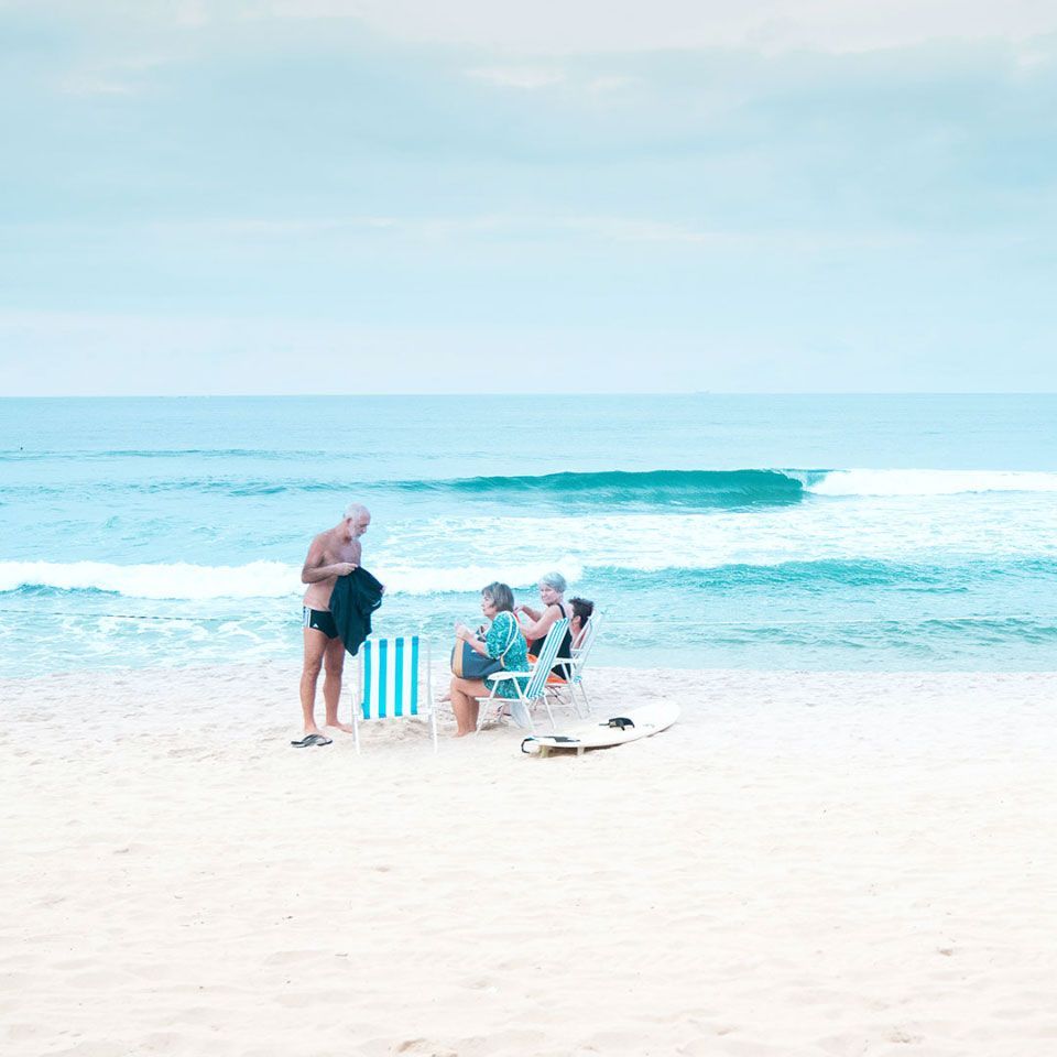 Beach scene: People in beach chairs, one man changing near the water. Blue sky, white sand, and ocean.