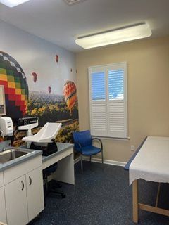 Doctor's office exam room with colorful hot air balloon mural. Exam tables, sink, and window with shutters are visible.