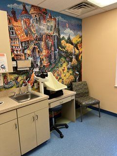 Pediatrician's office examination room with whimsical mural, weight scale, sink, cabinets, desk, and chair.