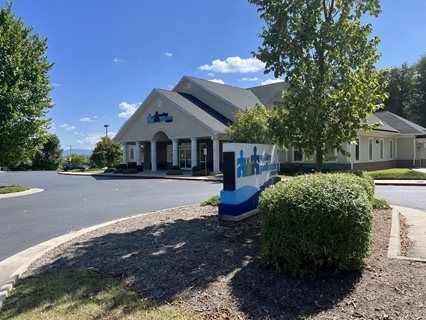 Office building with a blue and white sign and a small bush in front. Sunny day.