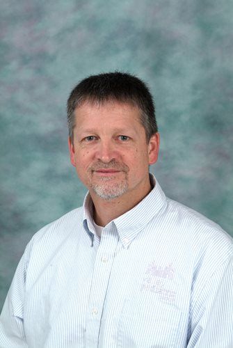 Man with short hair and goatee in a light blue striped shirt, smiling in front of a mottled blue-green background.