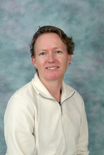 Woman with short brown hair, wearing a cream-colored zip-up jacket, smiling in front of a blue and green background.