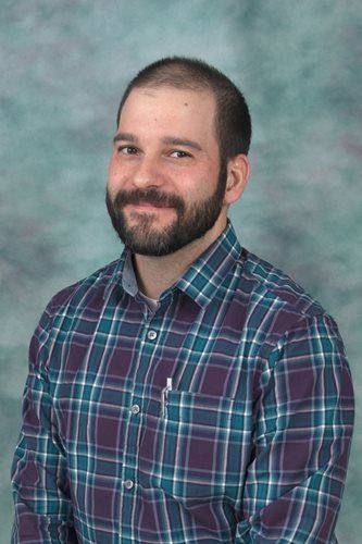 Man with beard wearing a plaid shirt, smiling, standing in front of a blurry blue and green background.