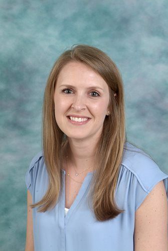 Woman with long blonde hair, smiling, wearing a light blue blouse, in front of a blue mottled background.