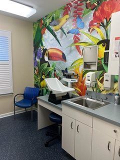 Pediatric examination room with a colorful jungle mural. Features a scale, sink, cabinets, and a blue chair.
