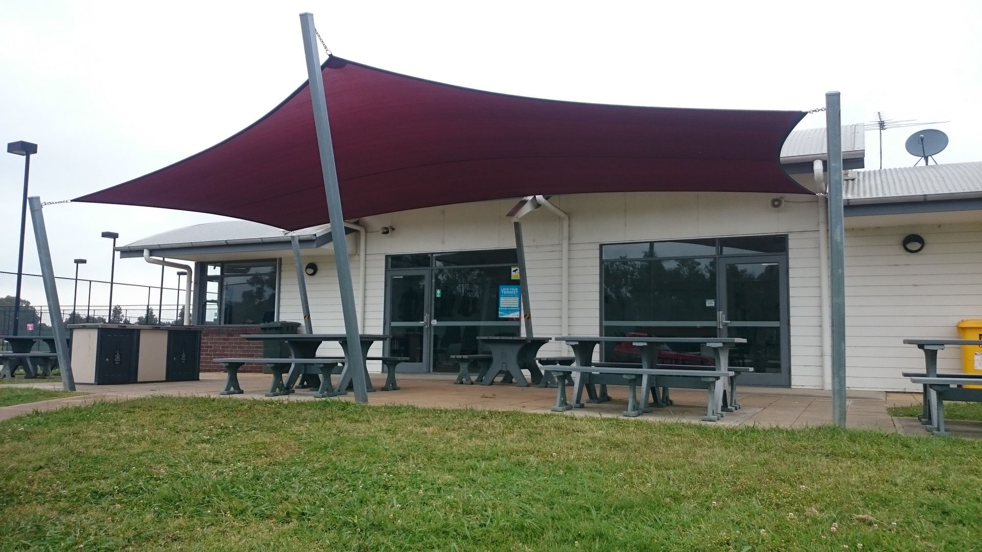 Custom maroon shade sail installed at Berwick Tennis Club, providing UV protection for spectator seating and enhancing the visual appeal and functionality of the club room, with sail tensioned over picnic tables in a landscaped outdoor area