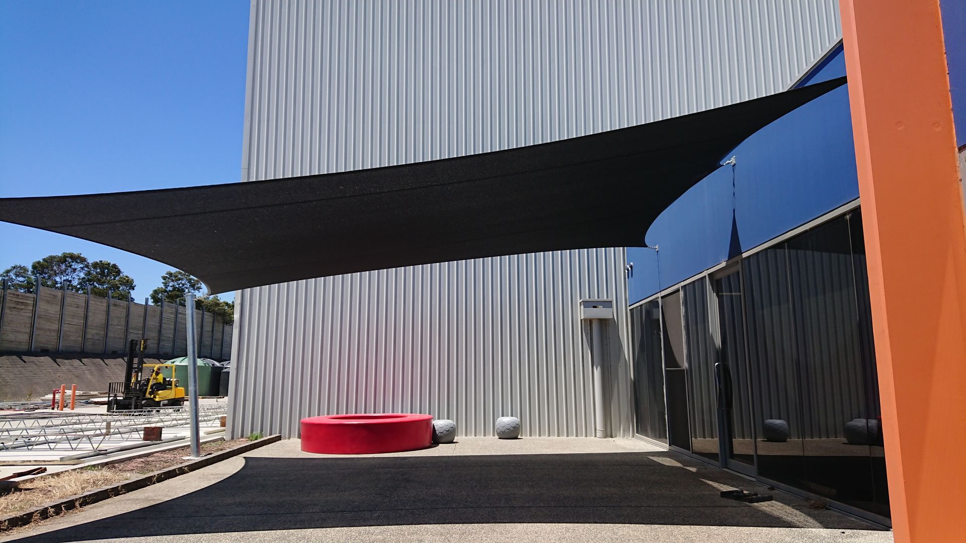 Industrial shade sail installation in Pakenham—custom black canopy tensioned between steel posts and a factory façade, creating a sheltered staff seating area in a new commercial development with red benches, stone seating, and enhancing the surrounding space and business 